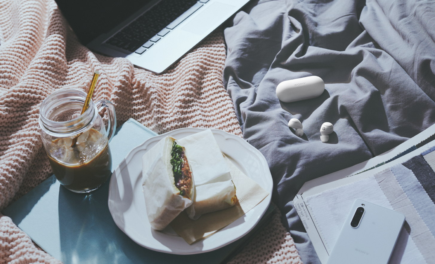 Image des écouteurs WF-C700N en blanc et de leur boîtier sur une nappe de pique-nique avec de la nourriture, des boissons, un ordinateur portable et un téléphone portable