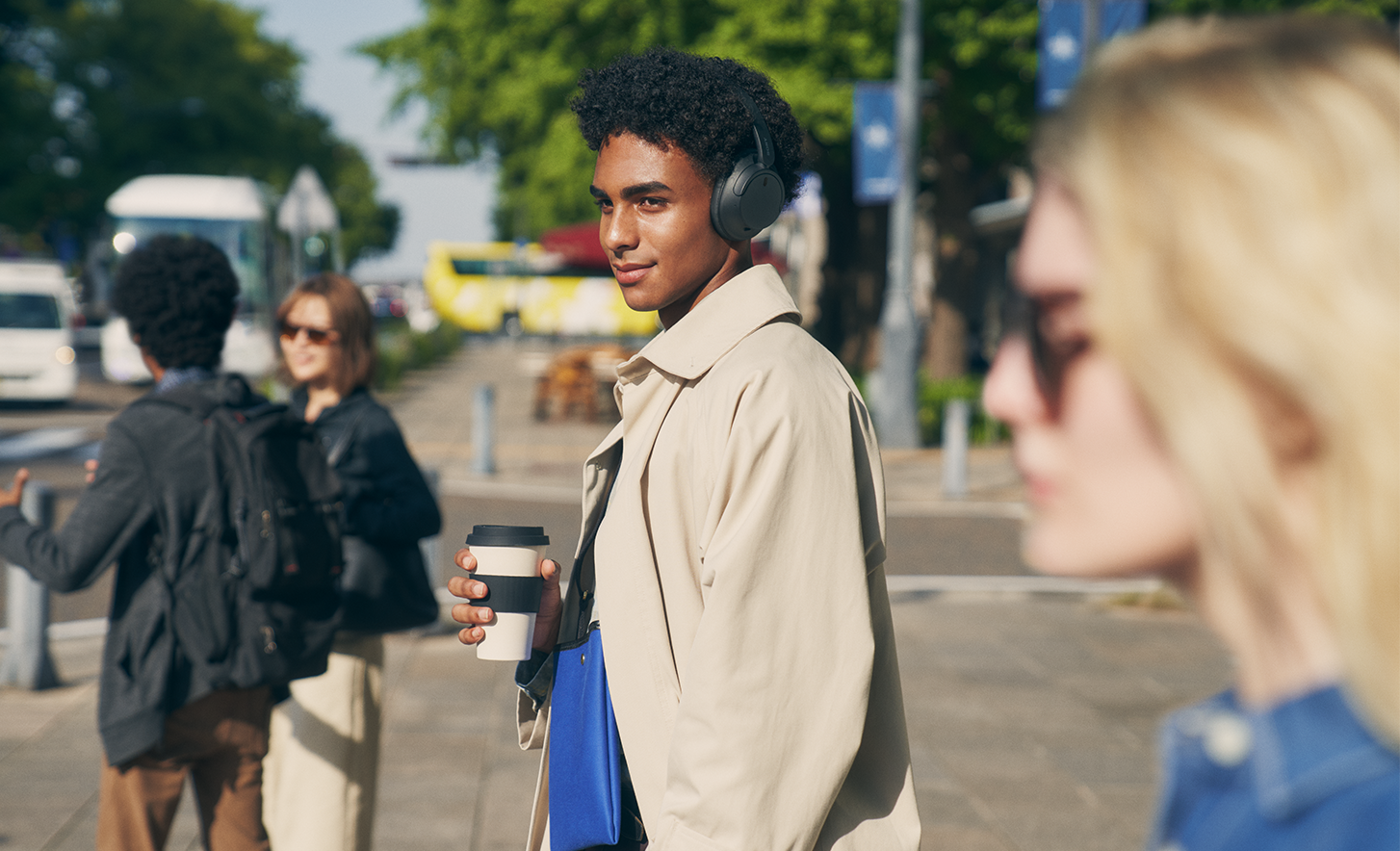 Image d'une personne dans une ville portant un casque Sony WH-CH720N noir et tenant une tasse de café.